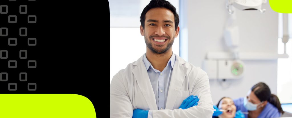 Um dentista de jaleco branco e luvas azuis sorri para a câmera; ao fundo, uma pessoa recebe atendimento odontológico de outro profissional.