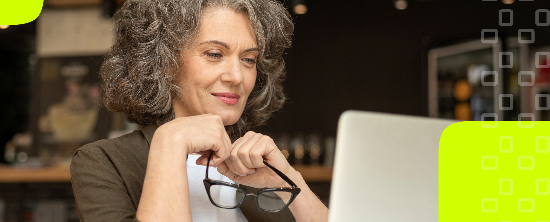Mulher de 40 anos com cabelo grisalho, as mãos próximas ao queixo segurando um óculos e olhando para o computador.